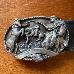 May include: Silver-toned metal belt buckle featuring a cowboy roping a horse, with two other horses and a cowboy figure. The buckle is oval-shaped and attached to a black leather belt. The design is detailed and textured.