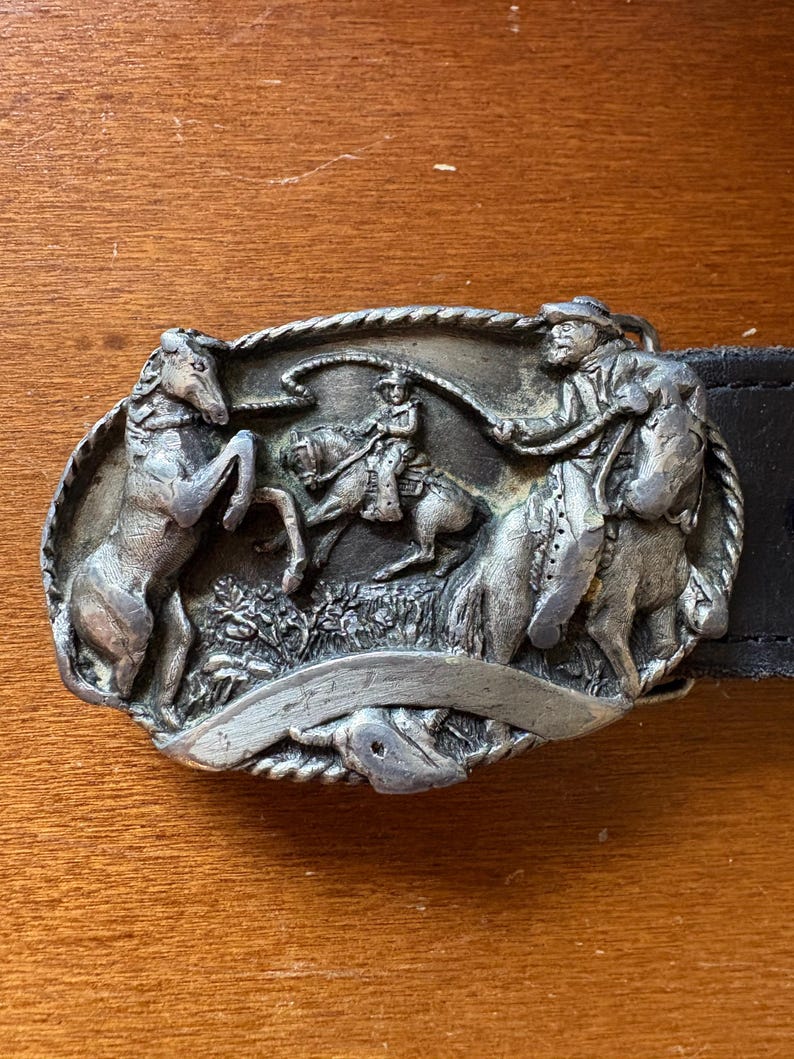 May include: Silver-toned metal belt buckle featuring a cowboy roping a horse, with two other horses and a cowboy figure. The buckle is oval-shaped and attached to a black leather belt. The design is detailed and textured.