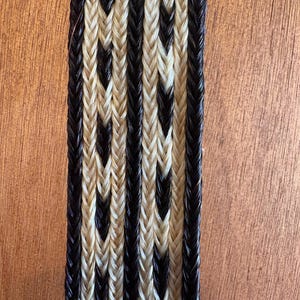 May include: Close-up of a woven strip with a repeating pattern of black and cream-colored braids. The braids are tightly woven together, creating a textured surface. The strip is likely a decorative element or part of a larger craft project.