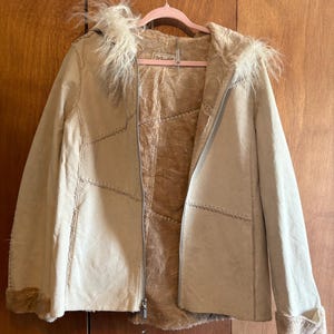 May include: A tan suede jacket with a faux fur-lined hood and cuffs. The jacket has a zipper closure and decorative stitching. The interior is lined with a lighter tan material. The jacket is hanging on a pink hanger.
