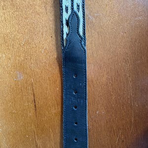 May include: A black leather belt with a woven cream and black pattern. The belt has several holes for adjustment and a decorative stitched design at the top. The belt is displayed against a wooden background.