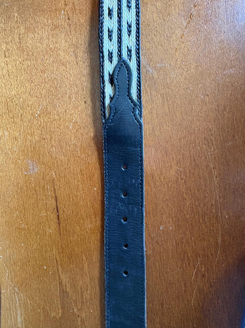 May include: A black leather belt with a woven cream and black pattern. The belt has several holes for adjustment and a decorative stitched design at the top. The belt is displayed against a wooden background.
