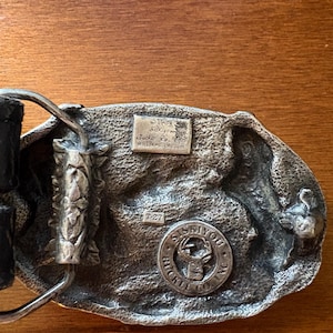 May include: A vintage silver-toned belt buckle with a detailed design. The buckle features a circular emblem with a deer and the text "SISKIYOU BUCKLE CO. INC." along with other text. The back of the buckle has additional text and the number "T-27".