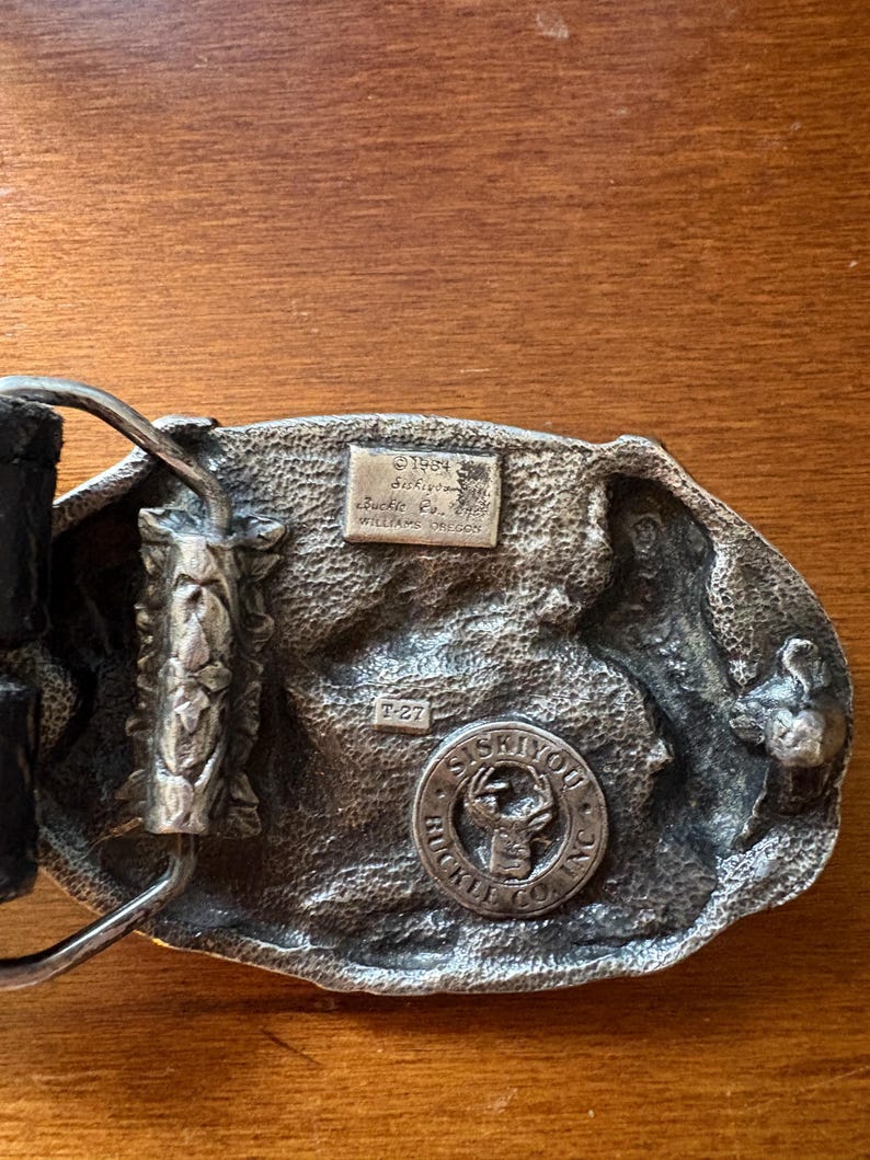 May include: A vintage silver-toned belt buckle with a detailed design. The buckle features a circular emblem with a deer and the text "SISKIYOU BUCKLE CO. INC." along with other text. The back of the buckle has additional text and the number "T-27".