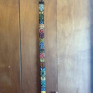 May include: A long, colorful belt with a gold-colored buckle. The belt is decorated with a repeating pattern of stylized floral designs in various colors, including green, yellow, pink, and blue. The belt is hanging vertically.