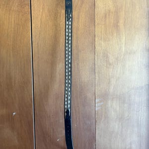 May include: Black leather belt with a silver buckle featuring an engraved design. The belt has a woven pattern in the center with black and beige threads. The belt is hanging vertically against a wooden background.