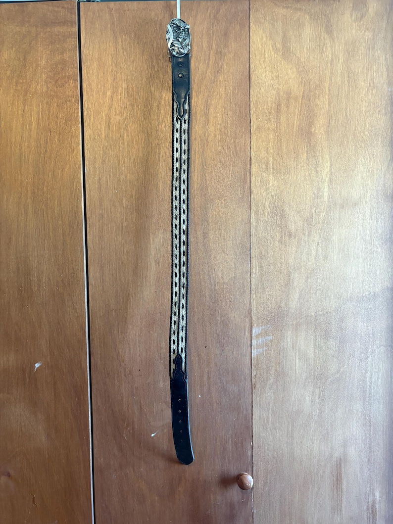 May include: Black leather belt with a silver buckle featuring an engraved design. The belt has a woven pattern in the center with black and beige threads. The belt is hanging vertically against a wooden background.