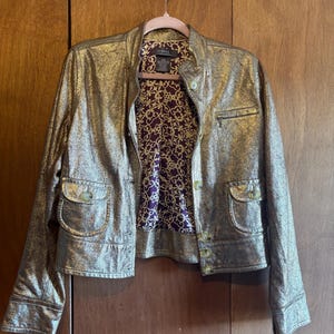 May include: Gold metallic jacket with a stand-up collar, two front pockets, and button closures. The jacket's interior lining features a purple and gold floral pattern. The jacket is hanging on a pink hanger.