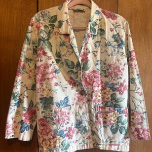 May include: A cream-colored blazer with a vibrant floral pattern featuring pink roses, blue hydrangeas, and yellow accents. The jacket has two front pockets and a single button closure. The blazer is hanging on a pink hanger.