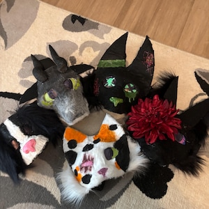 May include: Five handcrafted animal masks featuring various designs: a grey horned creature, a black cat with green accents, a white calico cat with orange and black spots, a black mask with a large red flower, and a black and white furry mask.