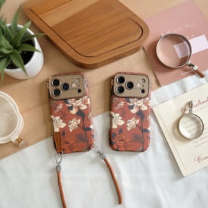 May include: Two phone cases with a floral pattern in shades of red, cream, and brown. Each case has a brown leather strap. The cases are on a light brown surface, with a wooden cutting board and a small plant in the background.