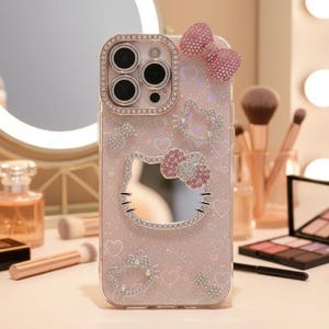 May include: A pink phone case with a Hello Kitty design, embellished with rhinestones. The case features a bow and heart accents, and a mirrored Hello Kitty face. The camera lens is surrounded by a rhinestone border.