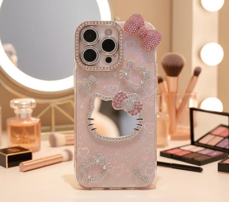 May include: A pink phone case with a Hello Kitty design, embellished with rhinestones. The case features a bow and heart accents, and a mirrored Hello Kitty face. The camera lens is surrounded by a rhinestone border.