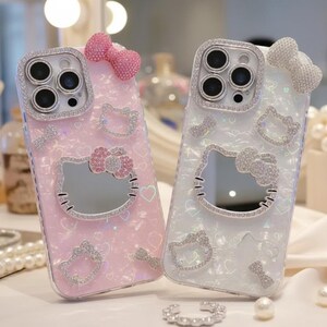 May include: Two decorative phone cases. One is pink, the other is white. Both cases feature a Hello Kitty design with a mirror, rhinestones, and a bow. The cases have a clear, sparkly finish and are embellished with hearts.