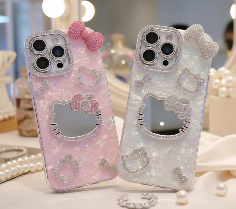 May include: Two decorative phone cases. One is pink, the other is white. Both cases feature a Hello Kitty design with a mirror, rhinestones, and a bow. The cases have a clear, sparkly finish and are embellished with hearts.