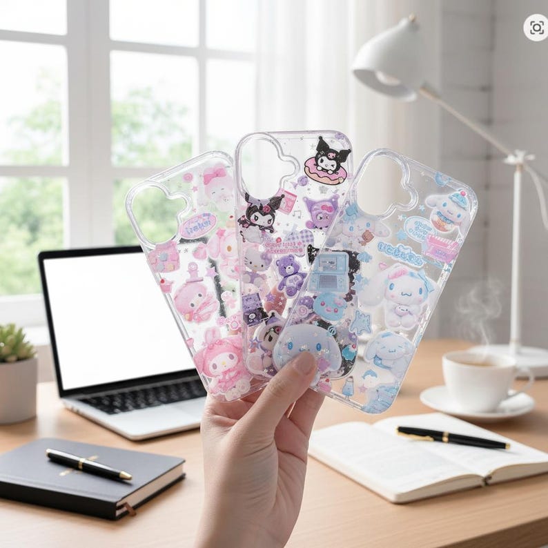 May include: Three clear phone cases with colorful cartoon character designs. The cases feature various characters in pink, purple, and blue, with decorative elements like bows, hearts, and stars. The cases are held in front of a desk with a laptop and notebook.