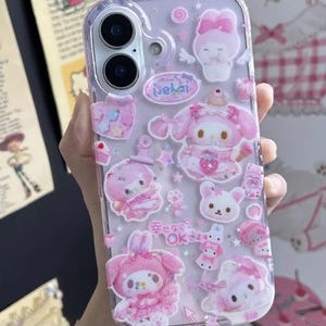 May include: Clear phone case decorated with pink and white cartoon characters and Japanese text. The case features a variety of designs, including bunnies, bows, and stars. The case is designed to fit a smartphone.