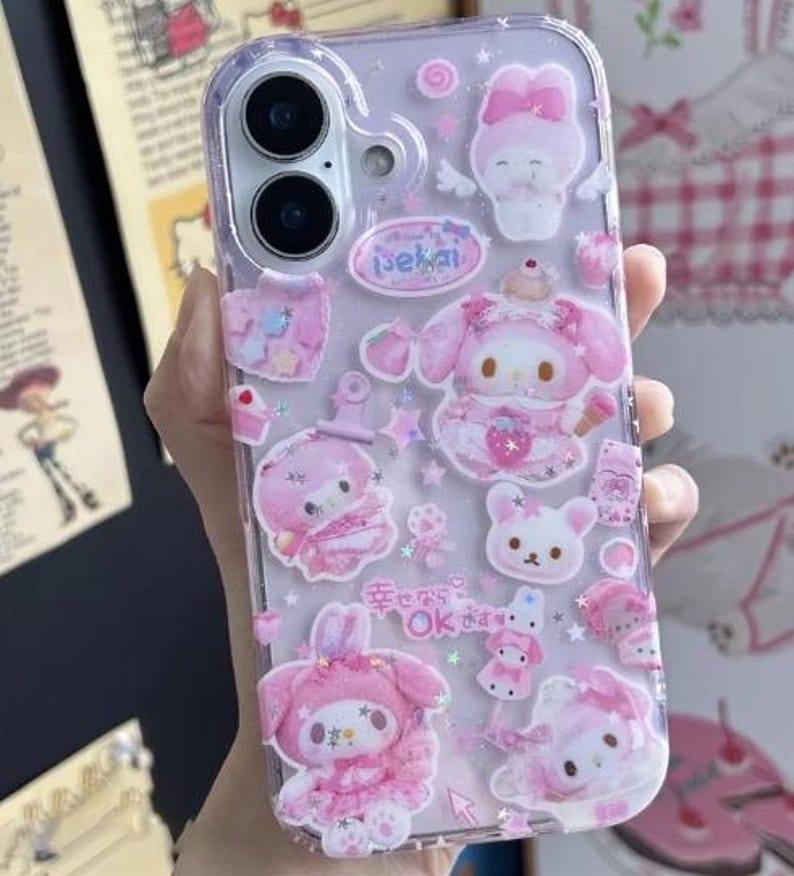 May include: Clear phone case decorated with pink and white cartoon characters and Japanese text. The case features a variety of designs, including bunnies, bows, and stars. The case is designed to fit a smartphone.