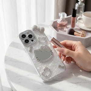 May include: A clear phone case with a sparkling design featuring a Hello Kitty motif. The case is adorned with rhinestones around the camera and on the Hello Kitty design, bows, and other shapes. The case is on a white marble table.