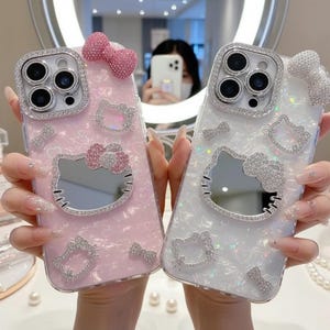 May include: Two decorative phone cases, one pink and one white, each featuring a Hello Kitty design. The cases are embellished with sparkling rhinestones and bows. The pink case has a pink bow and the white case has a silver bow.