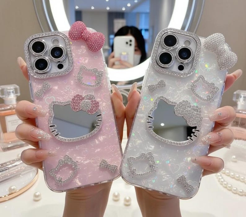 May include: Two decorative phone cases, one pink and one white, each featuring a Hello Kitty design. The cases are embellished with sparkling rhinestones and bows. The pink case has a pink bow and the white case has a silver bow.
