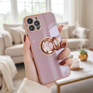 May include: A lavender phone case with a gold-colored ring holder. The case has a gold-colored border around the camera and edges. The ring holder is circular and has a metallic finish. The phone case is held in a hand.
