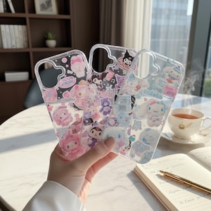 May include: Three clear phone cases with colorful cartoon character designs. The cases feature characters in pink, purple, and blue, with various shapes and patterns. The cases are held in front of a blurred background with a cup of tea and a notebook.