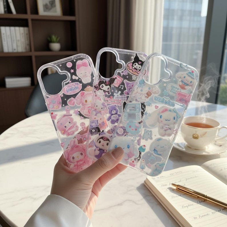 May include: Three clear phone cases with colorful cartoon character designs. The cases feature characters in pink, purple, and blue, with various shapes and patterns. The cases are held in front of a blurred background with a cup of tea and a notebook.