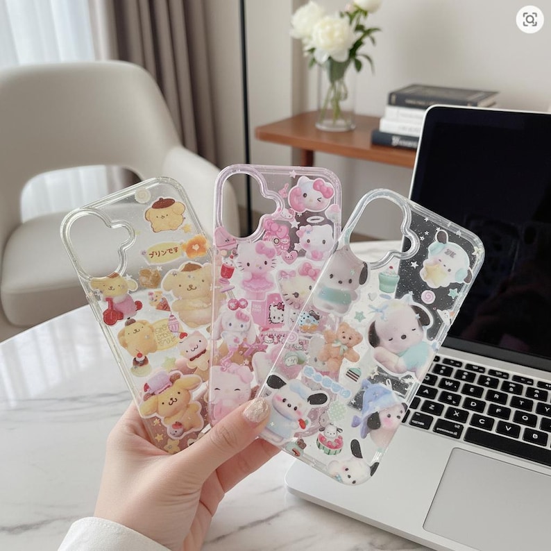 May include: Three clear phone cases with colorful cartoon character designs. One case features yellow characters with Japanese text, another has pink Hello Kitty designs, and the third showcases various characters. The cases are held in front of a laptop.