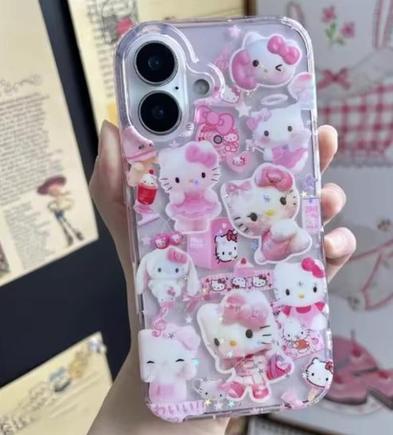 May include: Clear phone case with multiple pink Hello Kitty and other character designs. The case has a variety of 3D embellishments, including bows, dresses, and other decorative elements. The case is designed to fit a smartphone.