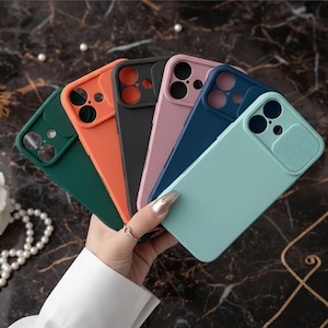 Puede incluir: Una mano sostiene un abanico de fundas de teléfono coloridas. Las fundas son verdes, naranjas, negras, rosas, azules y celeste. Cada funda tiene un protector de lente de cámara. El fondo es una superficie oscura, similar al mármol.