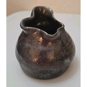 May include: A small, dark brown ceramic pitcher with a unique, organic shape. The pitcher has a wide, irregular opening and a textured surface. The glaze has a metallic sheen, reflecting light. The overall design is rustic and handcrafted.