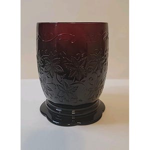 May include: A dark burgundy glass tumbler with a floral design. The tumbler has a wide base and a cylindrical shape, with embossed flowers and vines. The glass is a deep, rich color, and the design is intricate and detailed.