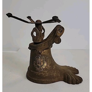 May include: An antique brass sprinkler head with a decorative footed base. The sprinkler has a central column with a rotating arm and a detailed emblem on the base. The metal has a weathered patina.