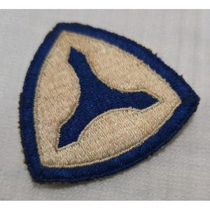 May include: A triangular embroidered patch featuring a dark blue border and a central dark blue three-pointed star shape set against a light beige background. The patch has a stitched edge and a textured surface created by dense thread work.