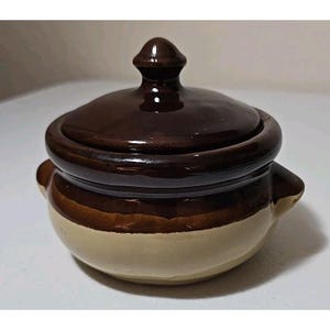 May include: A small, ceramic lidded pot with a dark brown lid and rim, a lighter brown band, and a cream-colored base. The pot has two small handles and a rounded shape. The lid has a small knob on top.