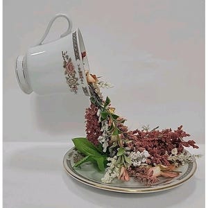 May include: A white teacup and saucer set with floral and geometric patterns. The teacup is tilted, spilling a cascade of dried flowers onto the saucer. The flowers are in shades of pink, white, and burgundy.