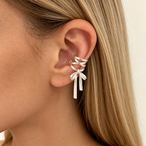 May include: Silver ear cuff featuring a bow design with dangling vertical bars. The ear cuff wraps around the ear and includes a spiral design. The jewelry is worn on the ear of a person with blonde hair.