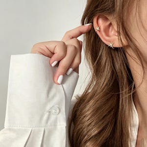 May include: Two gold-toned star-shaped earrings featuring small clear stones. One earring is positioned on the earlobe, while the other is worn as a cuff on the upper cartilage. The jewelry has a polished finish and a multi-pointed star design.