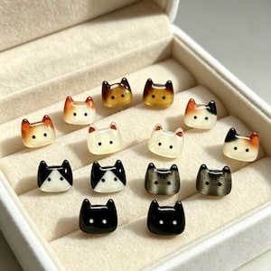 Puede incluir: Una colección de pequeños pendientes de botón con forma de cabeza de gato, con varios colores y patrones que incluyen diseños en negro sólido, blanco, calicó y carey. Cada pendiente tiene dos pequeños puntos para los ojos y orejas puntiagudas.
