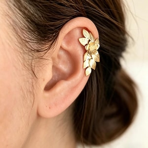 Puede incluir: Puño de oreja dorado con un diseño floral y de hojas. El puño se lleva en la oreja y tiene una estética delicada inspirada en la naturaleza. La pieza de joyería está hecha de metal y tiene un acabado pulido, añadiendo un toque de elegancia al aspecto general.
