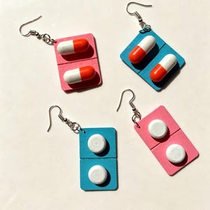 Puede incluir: Cuatro pares de pendientes originales. Dos pares presentan cuentas en forma de cápsula blancas y naranjas sobre respaldos rectangulares rosas y azules. Los otros dos pares tienen cuentas circulares blancas sobre respaldos rectangulares rosas y azules.