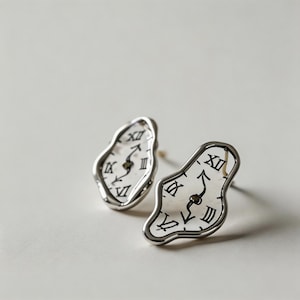 May include: A pair of silver-toned earrings shaped like melting clocks, reminiscent of a surrealist painting. Each earring features a white face with black Roman numerals and hands, creating a unique and artistic accessory.