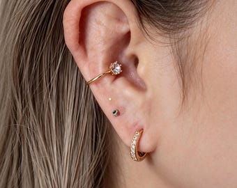 Könnte beinhalten: Ein goldfarbener Ear Cuff mit einem einzelnen klaren runden Stein, ein kleiner Ohrstecker mit einem winzigen grünen Stein und ein goldfarbener Creolen-Ohrring mit einer Reihe kleiner klarer Steine, getragen an Knorpel und Ohrläppchen.