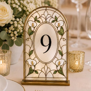 May include: An arched table number sign featuring the number 9 in black font centered within an oval. The frame is constructed with gold metal and translucent glass panels decorated with white calla lily flowers and green leaves.
