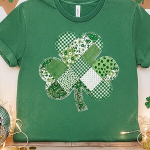 May include: Green t-shirt featuring a large shamrock design. The shamrock is made of various green patterned fabrics, including gingham, floral, and polka dots. The shirt is a solid green color and has short sleeves.