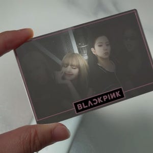Könnte beinhalten: Eine rechteckige Karte mit dem Text "BLACKPINK" in einem rosa und weißen Rechteck. Die Karte zeigt Bilder von vier Frauen. Die Karte wird vor einem weißen und grauen Hintergrund gehalten.