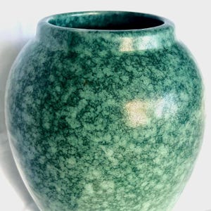 May include: A teal-coloured vase with a speckled pattern. The ceramic vase has a rounded body and a narrow opening. The surface has a mottled effect with light and dark teal hues. A decorative item.