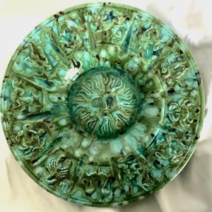 May include: A circular decorative plate with a detailed, raised design. The plate has a central sun-like motif and is mainly green with darker green and black accents. The rim features a repeating pattern of figures and shapes. The plate is likely ceramic.