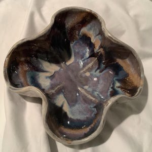 May include: A handcrafted ceramic bowl with a distinctive four-lobed design. The bowl is finished with a glossy glaze in shades of blue, purple, and tan. The interior showcases a floral pattern. This bowl is a decorative item.